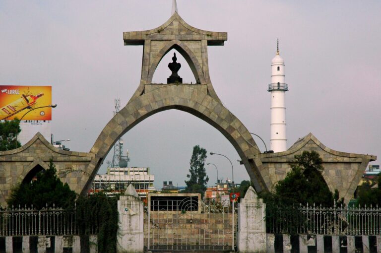 A photo showing a unique archway with a silhouetted monument at the center, adjacent to a white lighthouse against an overcast sky, with urban surroundings and a billboard to the left.