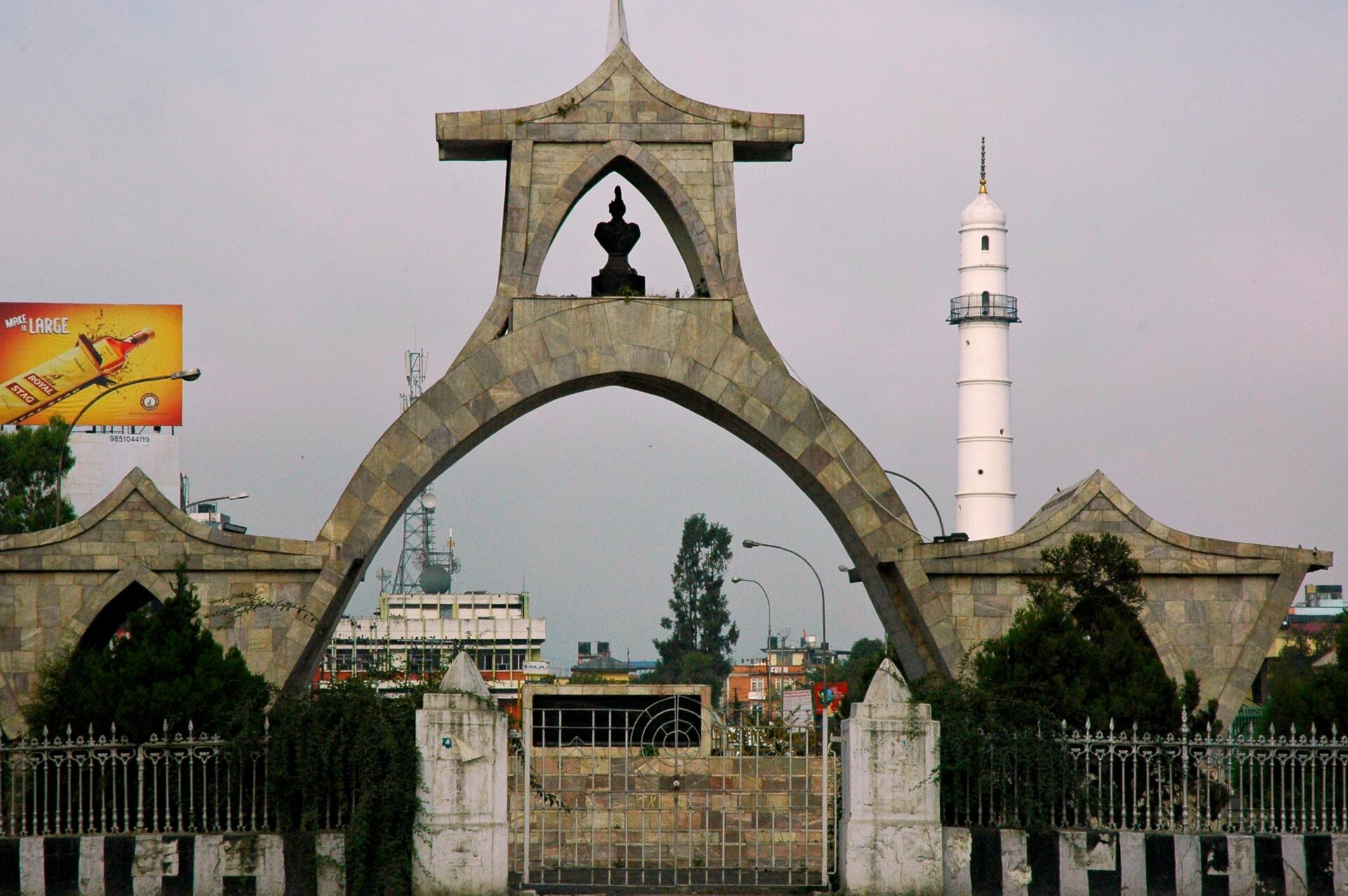 A photo showing a unique archway with a silhouetted monument at the center, adjacent to a white lighthouse against an overcast sky, with urban surroundings and a billboard to the left.