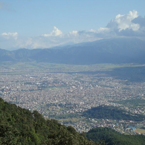 A panoramic view overlooking a densely populated city nestled in a valley with mountains in the background under a partly cloudy sky. Vegetation can be seen in the foreground.