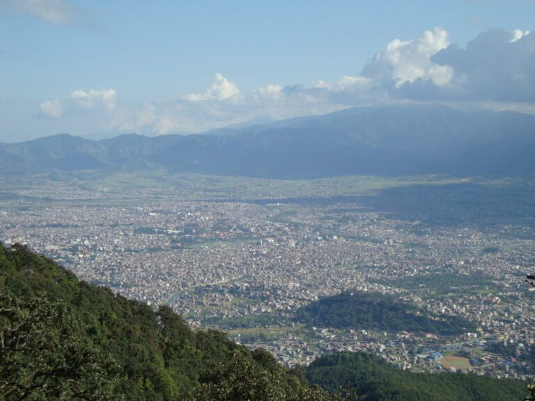 A panoramic view overlooking a densely populated city nestled in a valley with mountains in the background under a partly cloudy sky. Vegetation can be seen in the foreground.