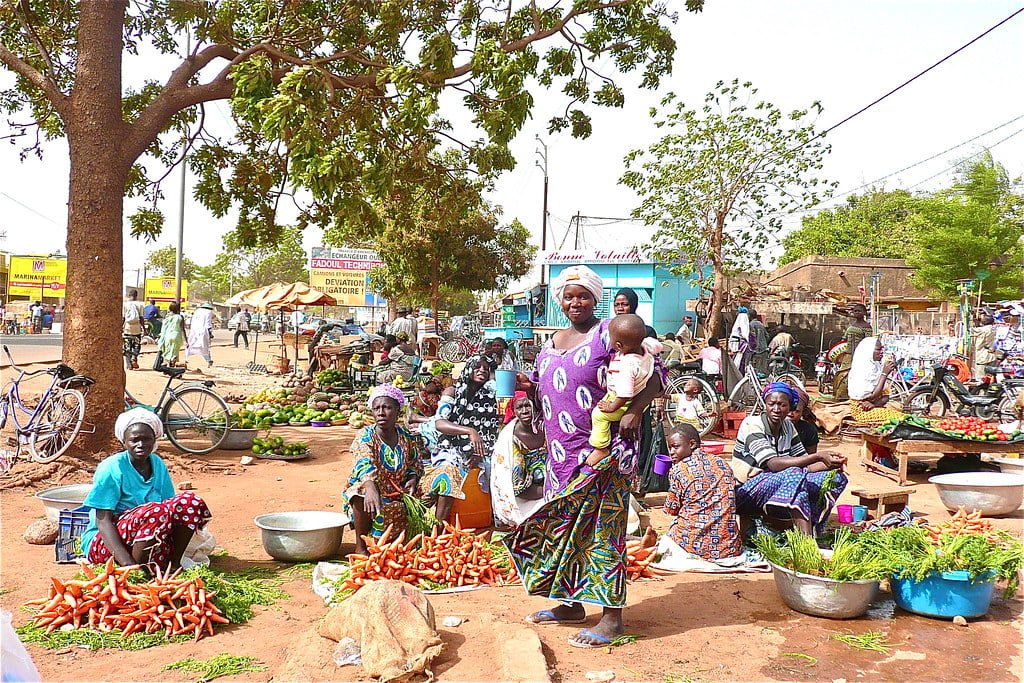 A vibrant street market scene in Africa with people selling vegetables and grains while a woman in brightly colored traditional clothing holds a child, with bicycles and motorbikes in the background.