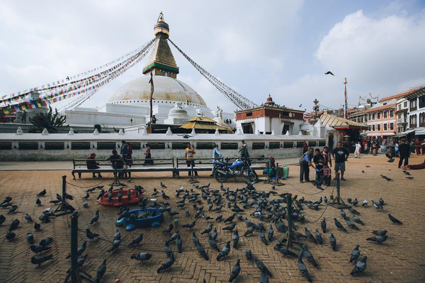 A bustling scene at Boudhanath Stupa in Kathmandu, Nepal, with prayer flags, pigeons congregating on the ground, and people walking and sitting around the perimeter.