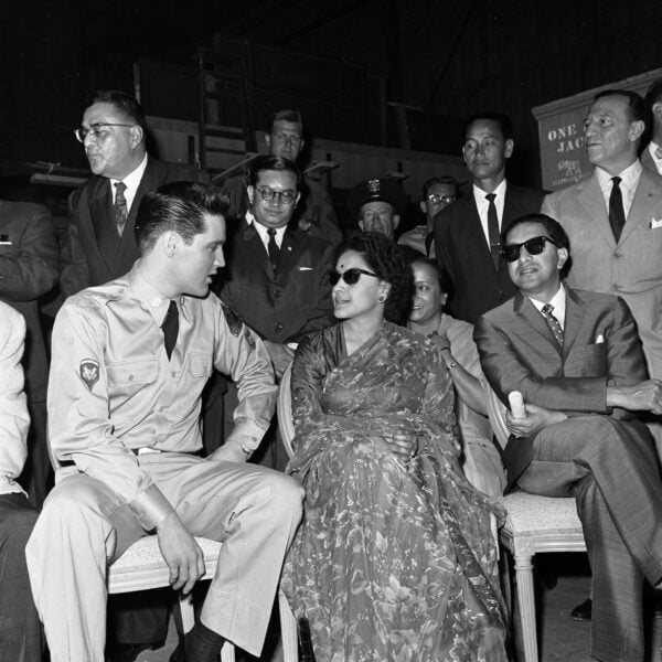 A black and white photo shows a young man in a military uniform seated and turned towards a woman in a floral dress and sunglasses. They are surrounded by a group of men in suits and onlookers in various attire.