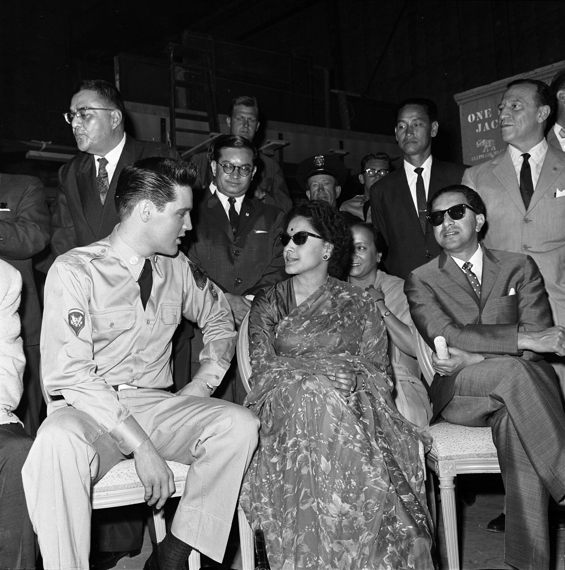 A black and white photo shows a young man in a military uniform seated and turned towards a woman in a floral dress and sunglasses. They are surrounded by a group of men in suits and onlookers in various attire.