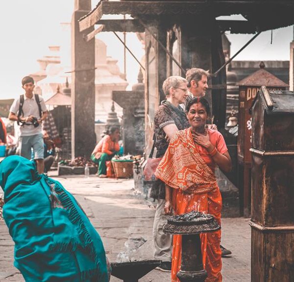 A woman in traditional orange attire smiling at the camera in a bustling street scene with other people and market stalls around her, some with smoke rising from street food cooking.