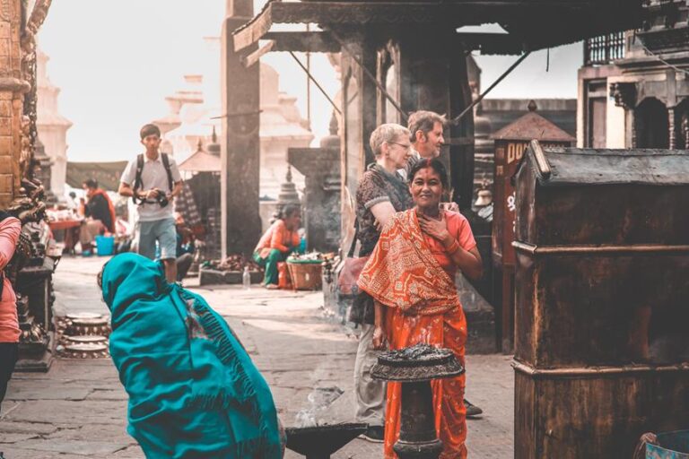 A woman in traditional orange attire smiling at the camera in a bustling street scene with other people and market stalls around her, some with smoke rising from street food cooking.