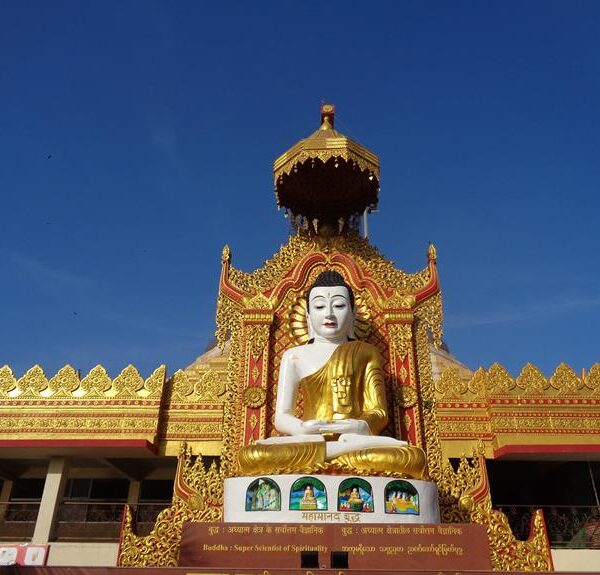 An ornate golden Buddha statue seated in meditation pose, sheltered by an intricate tiered roof with traditional patterns, against a blue sky. The statue is part of a temple structure adorned with golden embellishments and Burmese script.