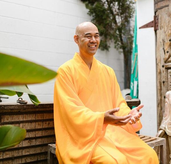 A smiling person in a yellow robe sitting next to a Buddha statue in a tranquil outdoor setting with green foliage and a carved wooden background.