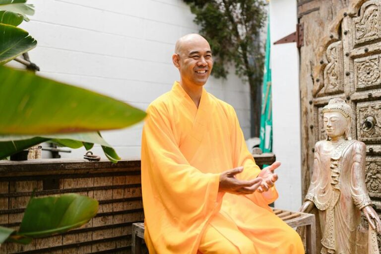 A smiling person in a yellow robe sitting next to a Buddha statue in a tranquil outdoor setting with green foliage and a carved wooden background.