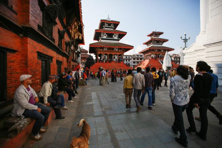 People walking and sitting around an open square with traditional multi-tiered buildings in the background and a dog in the foreground.