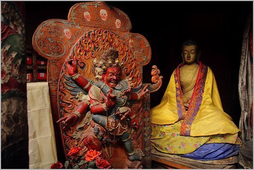 A colorful, ornate Buddhist shrine with a fearsome deity figure carved and painted in red next to a serene, golden-clad Buddha statue sitting in meditation.