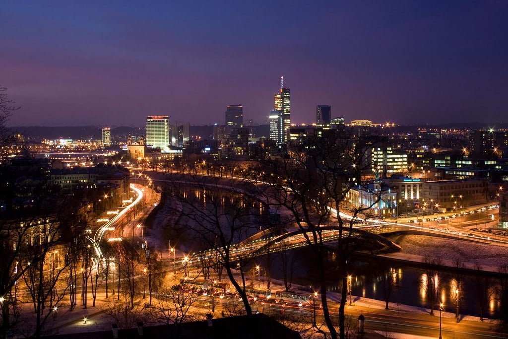 A nighttime cityscape with illuminated buildings and streets, featuring a riverfront and curving roads with light trails from moving vehicles.