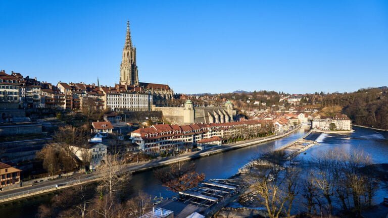 A panoramic view of a historic cityscape with prominent Gothic cathedral spire, traditional European buildings, a river flowing through, and clear blue skies.