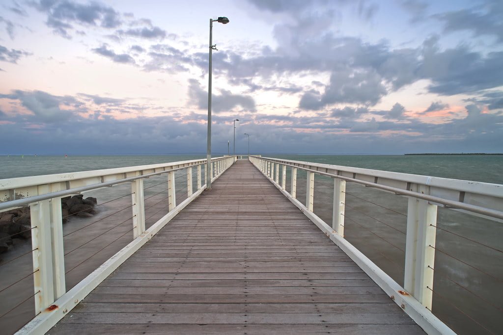 A wooden pier extends into the sea with a cloudy sky at dusk, featuring a lamp post and railing along its length.