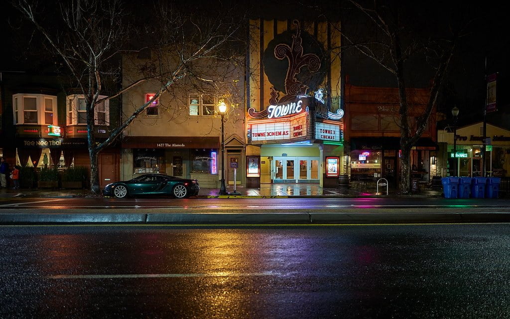 A night scene of a wet city street in front of a classic cinema with neon signs, its marquee lit up, and a dark-colored car parked at the curb.