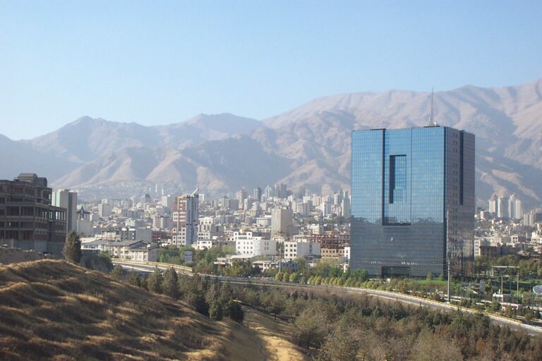 A cityscape with a mixture of modern and traditional buildings against a backdrop of hazy mountains, featuring a prominent glass skyscraper with a unique rectangular void in its center.