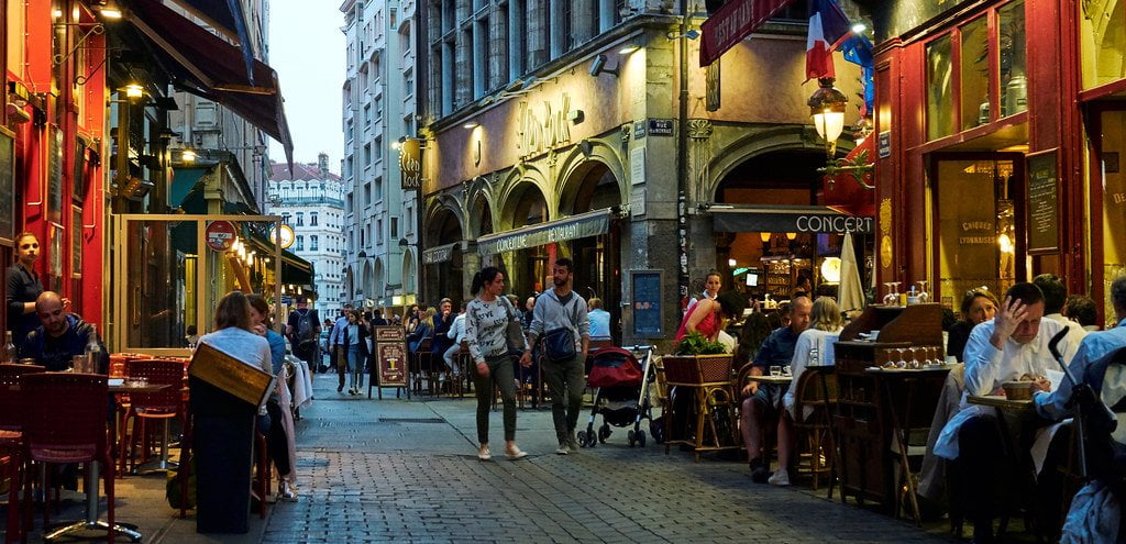 Alt text: A bustling evening street scene in a European city with people dining at outdoor restaurant tables and walking along the narrow cobblestone street flanked by colorful building facades.
