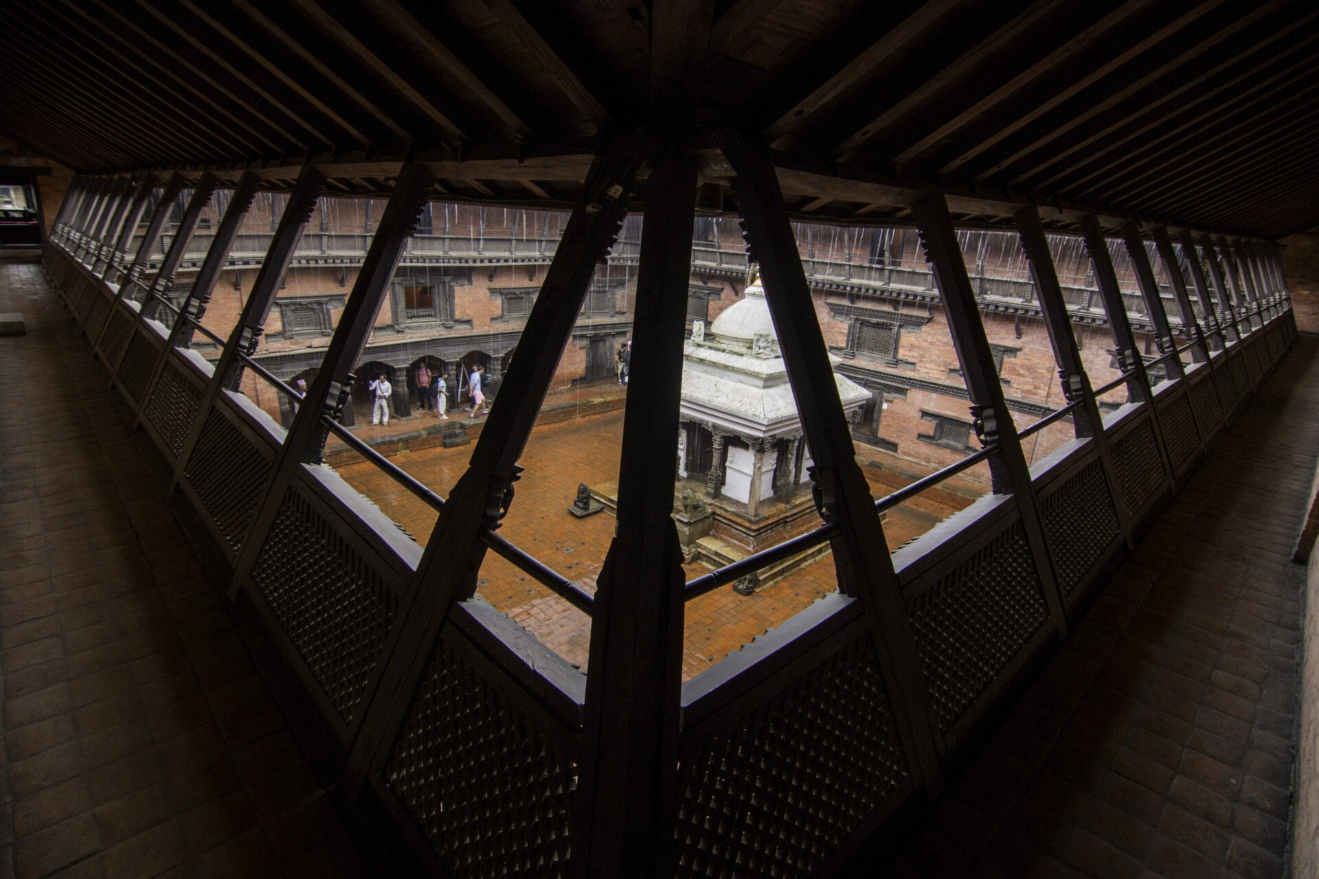 A view from within a dark wooden corridor with latticed windows overlooking a courtyard featuring traditional architecture and people gathered below.
