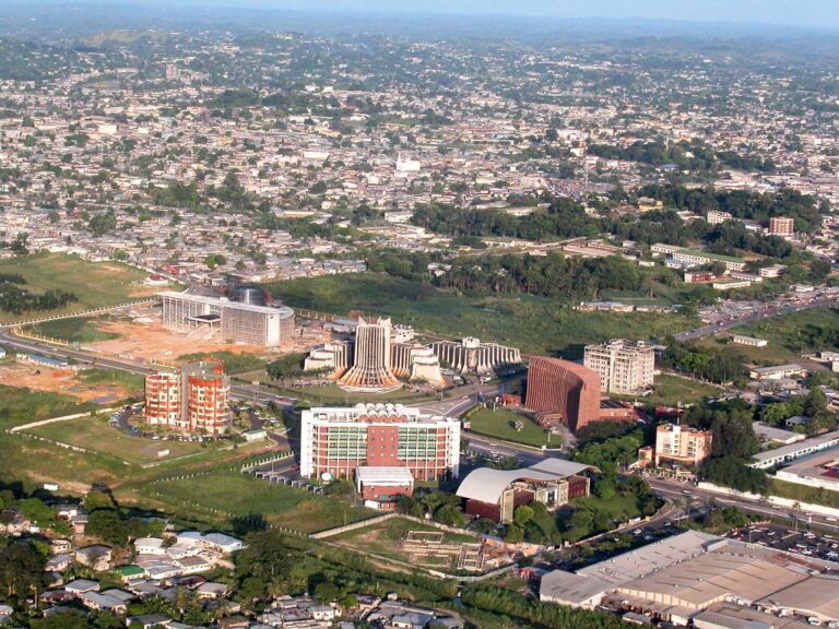 Aerial view of an expansive cityscape with dense housing, several mid-rise buildings under construction, a distinctive round building, and greenery interspersed throughout, under clear skies.