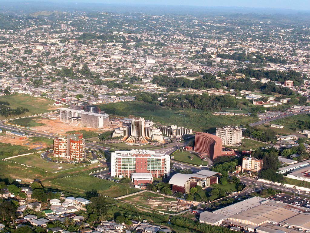Aerial view of an expansive cityscape with dense housing, several mid-rise buildings under construction, a distinctive round building, and greenery interspersed throughout, under clear skies.