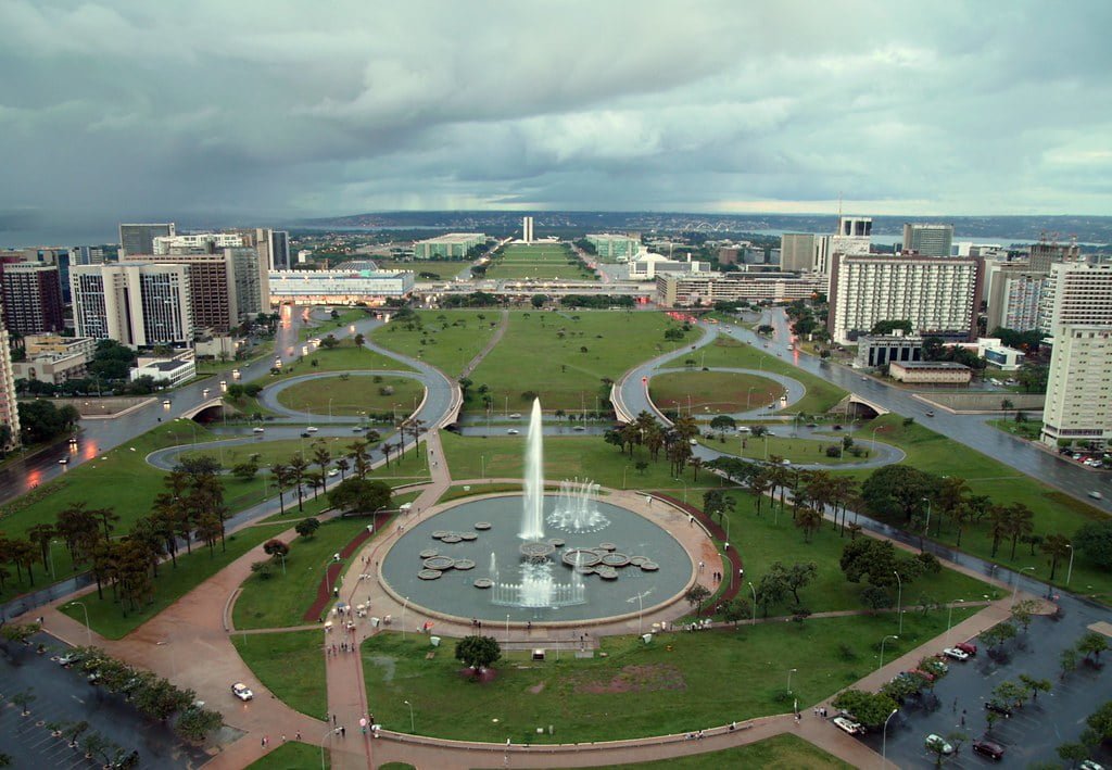 Aerial view of a cityscape featuring a large fountain in the foreground, with curving roads, lush green spaces, and buildings under a cloudy sky.