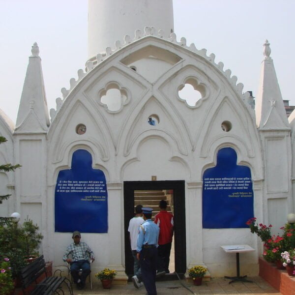 A white building with arched Gothic-style details and blue informational plaques on either side of the entrance where people are passing through. There are benches and potted flowers in the foreground.