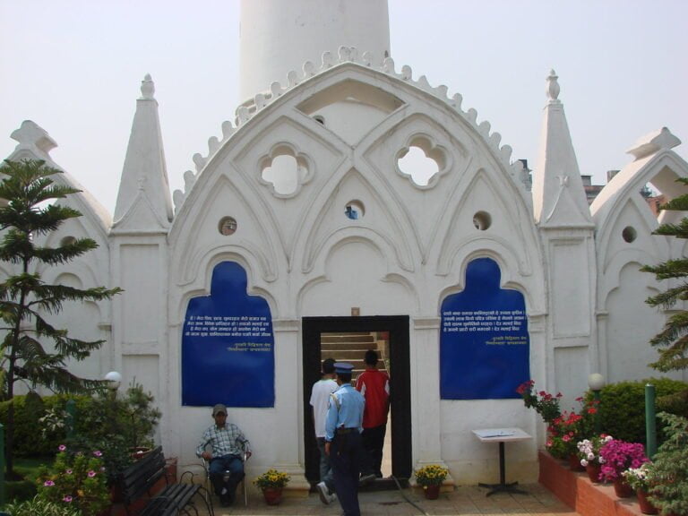 A white building with arched Gothic-style details and blue informational plaques on either side of the entrance where people are passing through. There are benches and potted flowers in the foreground.