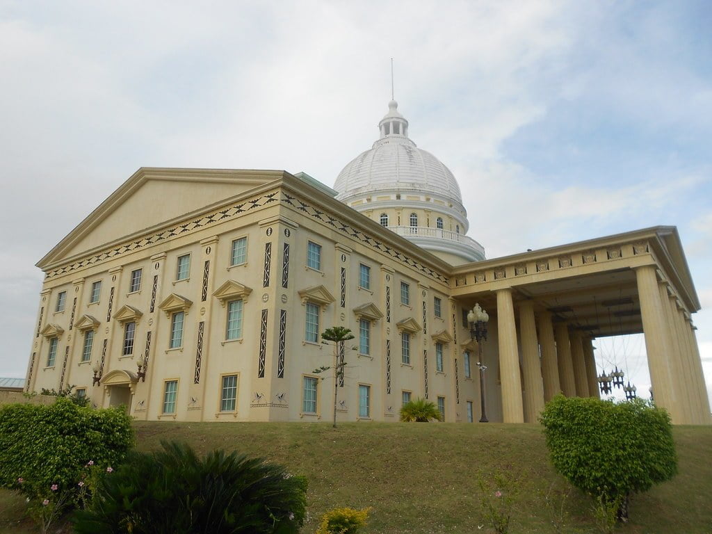 A view of a large neoclassical building with a dome and portico, located on a grassy hill with a few small shrubs in the foreground under a partly cloudy sky.