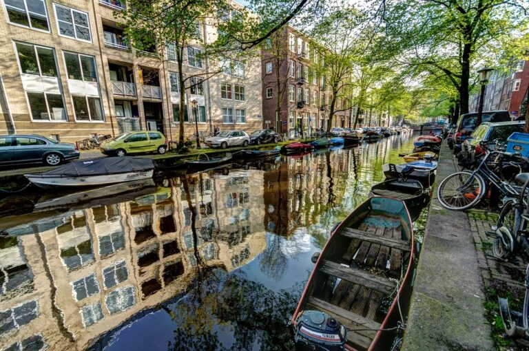 A canal in Amsterdam lined with cars, boats, and bicycles, with historical buildings reflected in the calm water under a canopy of green trees.