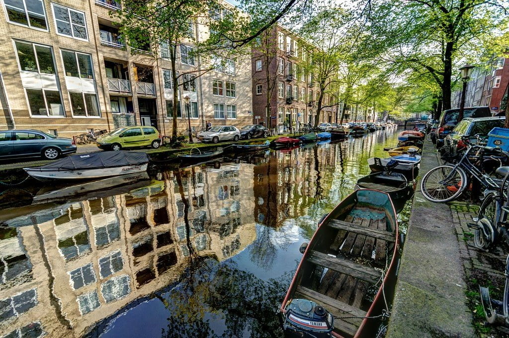 A canal in Amsterdam lined with cars, boats, and bicycles, with historical buildings reflected in the calm water under a canopy of green trees.