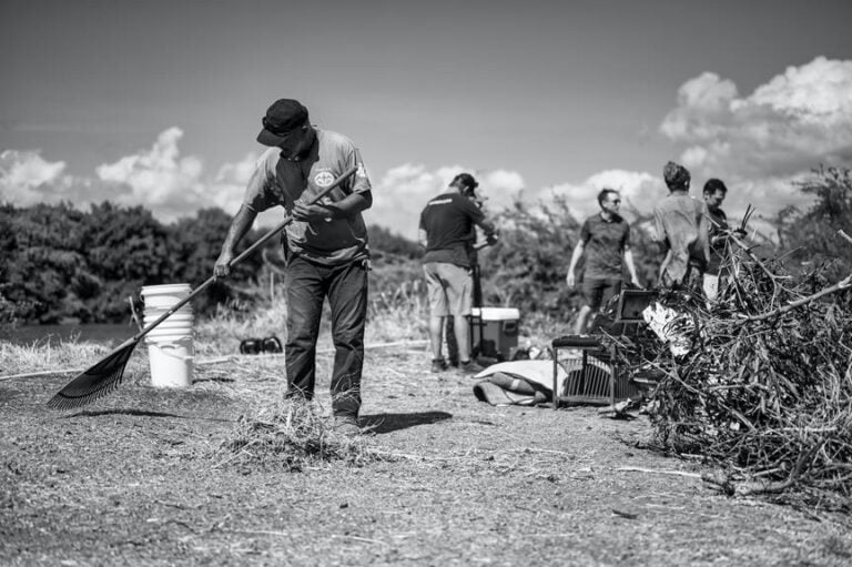 Black and white photo of a person raking the ground in the foreground, with several other people working on clearing brush and debris in the background, all under a cloudy sky.