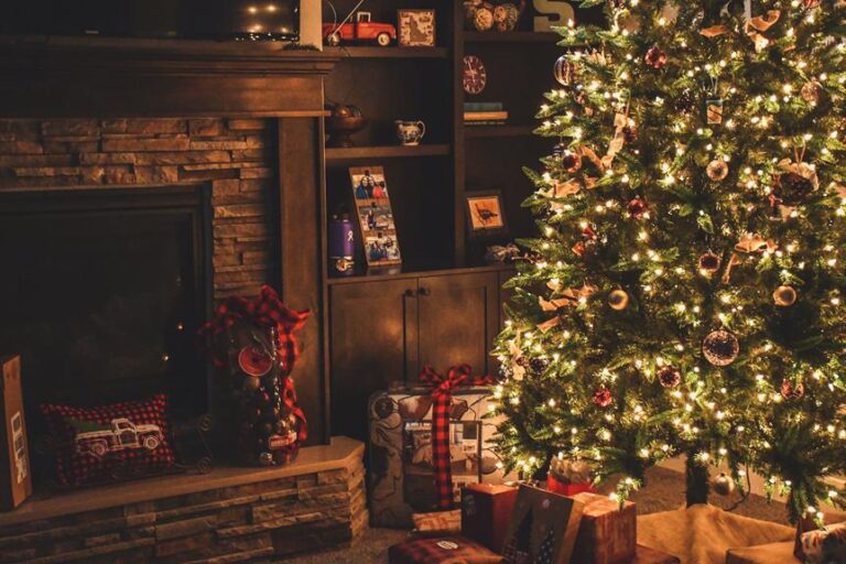 A warmly lit living room decorated for Christmas with a large, illuminated Christmas tree, presents underneath, a hearth with festive decorations, and a bookshelf filled with ornaments and photos.