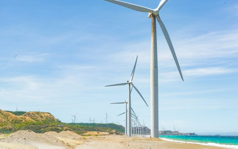 Row of large wind turbines at a coastal wind farm with clear blue sky, sandy beach in the foreground, and hilly terrain in the background.