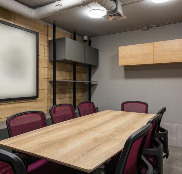 A modern meeting room with a large wooden table, red and black chairs, wood paneling on the wall, a blank framed picture, and shelving units.