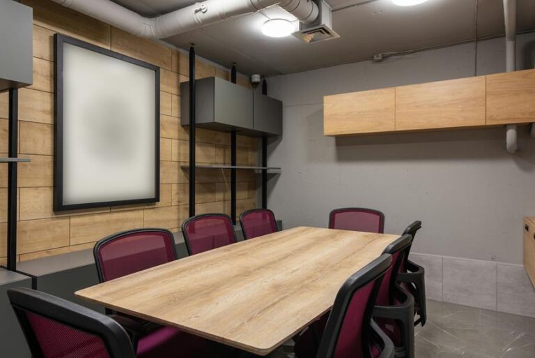 A modern meeting room with a large wooden table, red and black chairs, wood paneling on the wall, a blank framed picture, and shelving units.