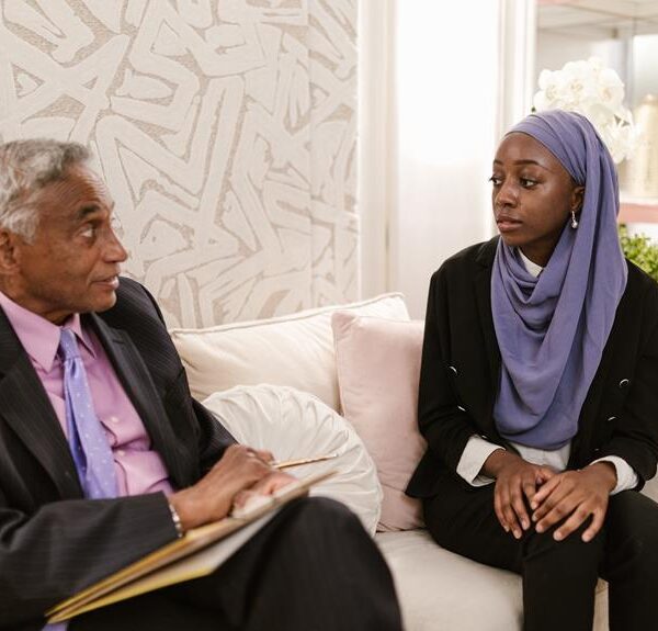 An elderly man in a suit holding a notebook sits facing a young woman in a hijab on a couch, appearing to be in a conversation in a well-lit room with elegant decor.