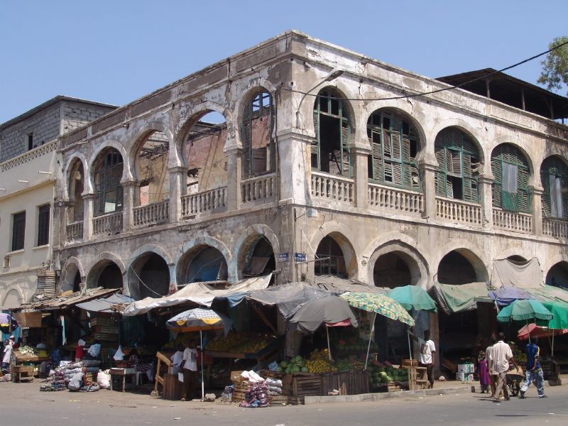 An old, weathered two-story building with arched windows and a balcony, sitting above a bustling street market with various colorful umbrellas and stalls in what appears to be an urban area.