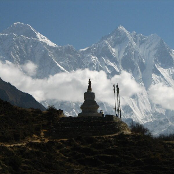 Alt text: A traditional stupa in the foreground with prayer flags stands against the backdrop of the snow-capped Himalayan mountains shrouded in clouds, with a clear blue sky above and a hiker visible on a trail.