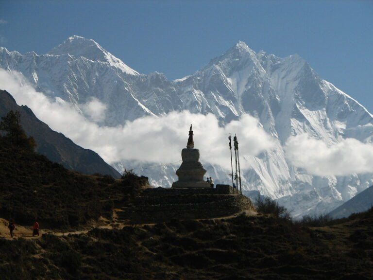 Alt text: A traditional stupa in the foreground with prayer flags stands against the backdrop of the snow-capped Himalayan mountains shrouded in clouds, with a clear blue sky above and a hiker visible on a trail.
