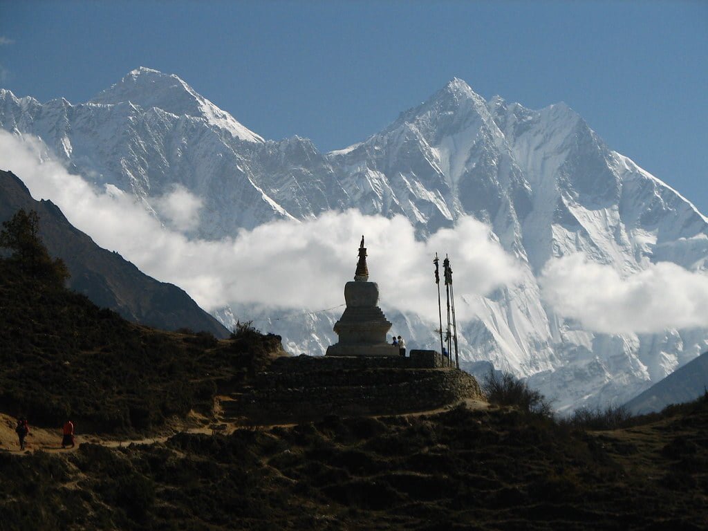 Alt text: A traditional stupa in the foreground with prayer flags stands against the backdrop of the snow-capped Himalayan mountains shrouded in clouds, with a clear blue sky above and a hiker visible on a trail.