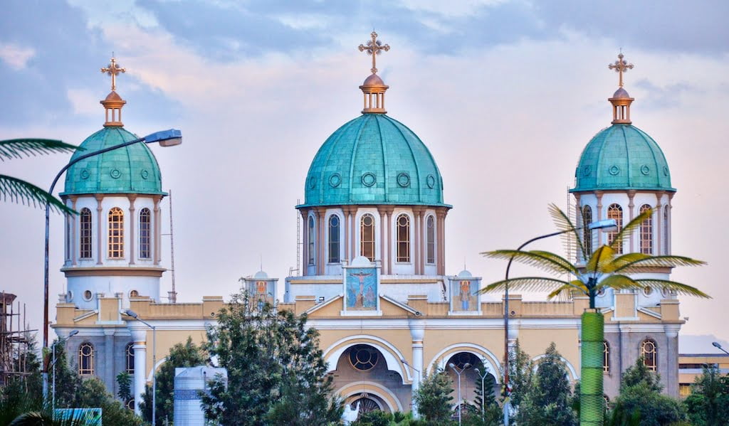 Alt text: A view of a church with two prominent turquoise domes topped with golden crosses, framed by palm trees against a dusky sky.