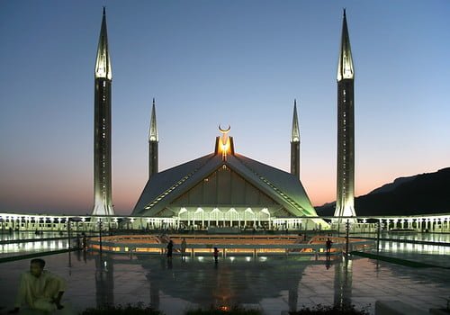 The Faisal Mosque in Islamabad, Pakistan, illuminated at twilight against a mountainous backdrop, with its modern design featuring a triangular prayer hall and four towering minarets. A reflection pool and a solitary figure in the foreground add to the tranquil scene.