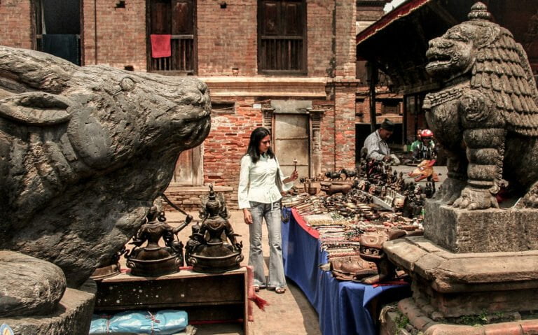 A woman browsing a street market with traditional wares displayed on tables, flanked by large stone statues of mythological creatures, set against a backdrop of old brick buildings.