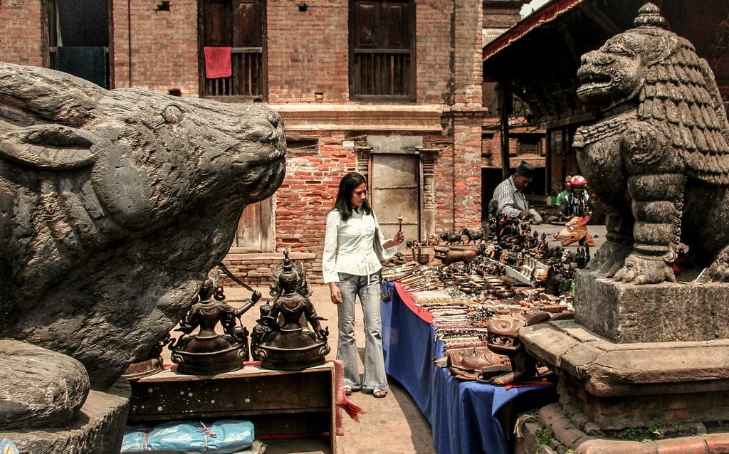 A woman browsing a street market with traditional wares displayed on tables, flanked by large stone statues of mythological creatures, set against a backdrop of old brick buildings.