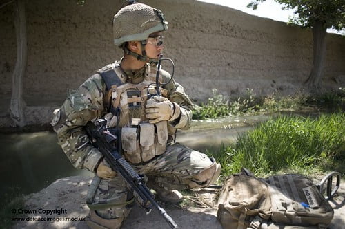 A soldier in camouflage gear and a helmet is kneeling beside a stream with a radio handset to their mouth, holding a rifle, with a backpack resting on the ground nearby.
