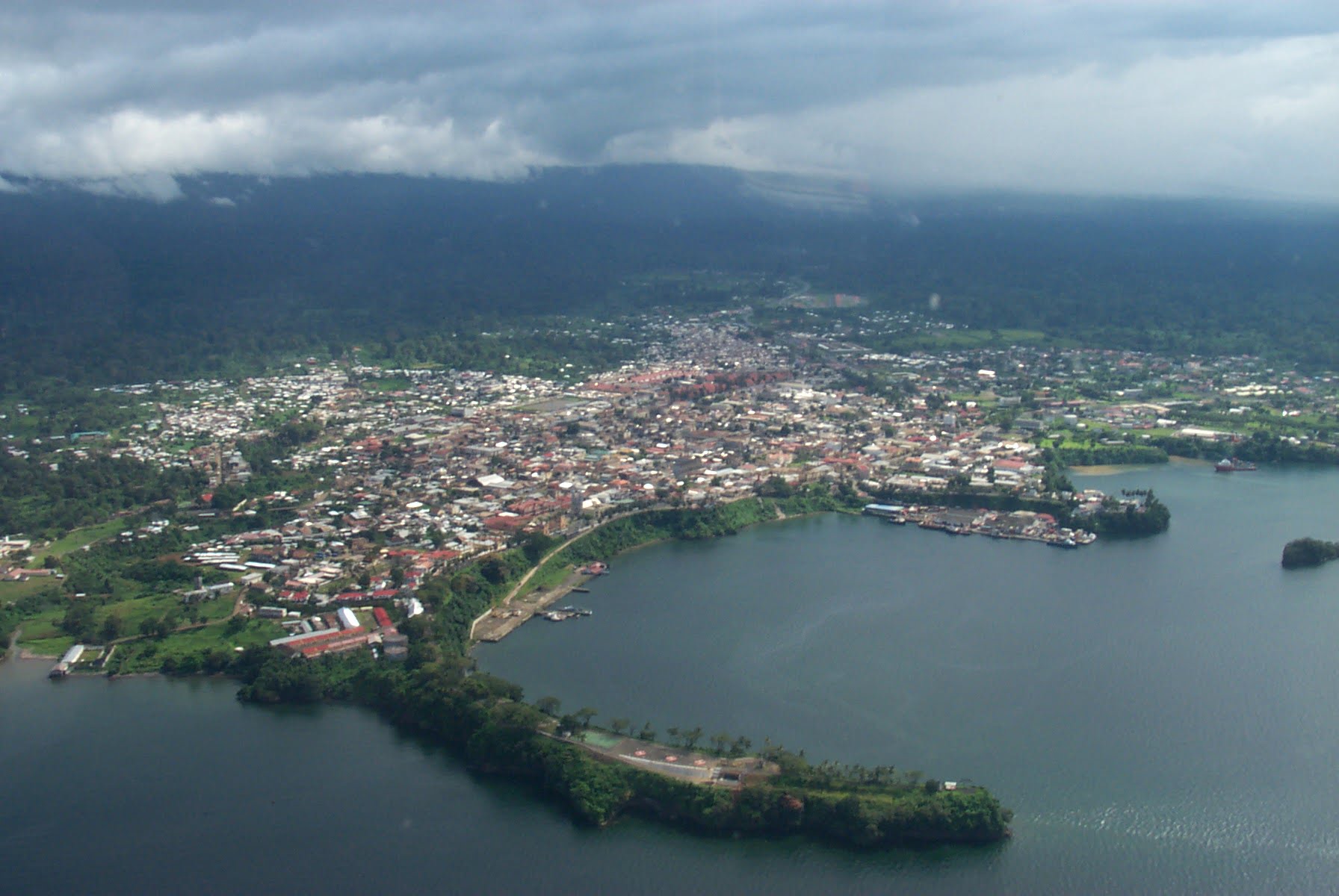 Aerial view of a coastal town with dense building clusters, bordered by a large body of water, under a cloudy sky.