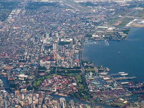 Aerial view of a densely populated coastal city with tall buildings, adjacent to a port with several ships, and a large airport runway in the vicinity.