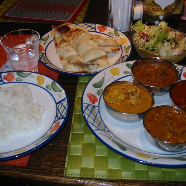 Alt text: A spread of Indian cuisine, including a plate of white rice, a tray with naan bread, a bowl of salad, and a plate with four small bowls of different curries, on a colorful table setting.
