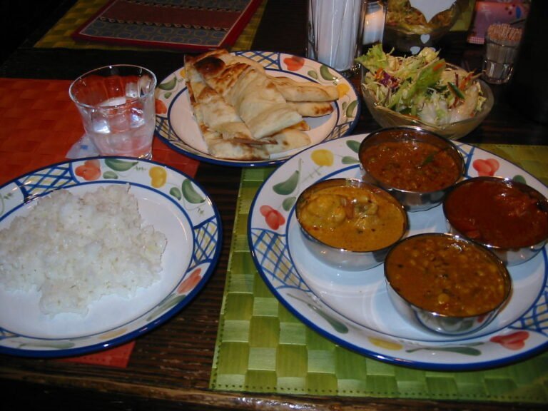 Alt text: A spread of Indian cuisine, including a plate of white rice, a tray with naan bread, a bowl of salad, and a plate with four small bowls of different curries, on a colorful table setting.