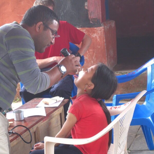 A man in a striped sweater is examining a young girl's eyes while she sits in a plastic chair, tilting her head back. They appear to be outdoors and part of a vision screening event.
